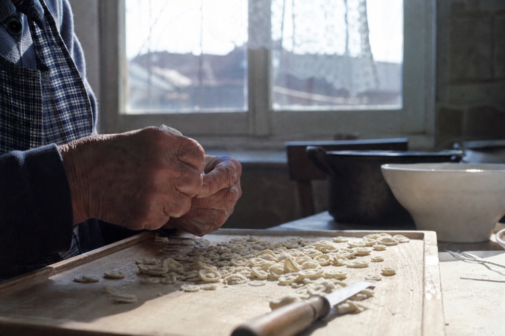 An older woman's weathered hands shaping small pasta pieces on a scratched wooden board in a plain village kitchen, a single window behind