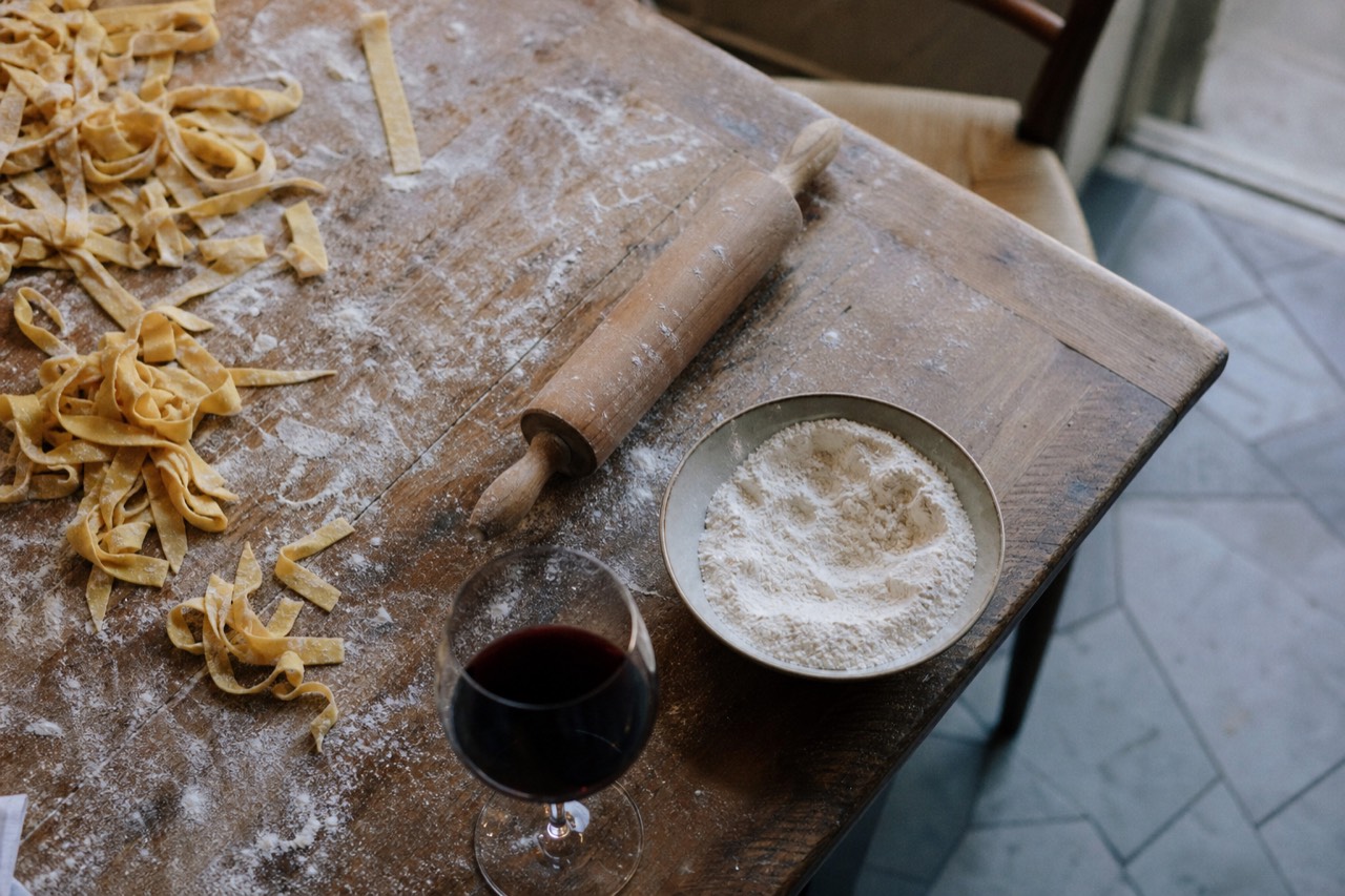 An overhead view of a floured wooden table with fresh egg pasta ribbons, a rolling pin, a bowl of flour and a glass of red wine in warm afternoon light