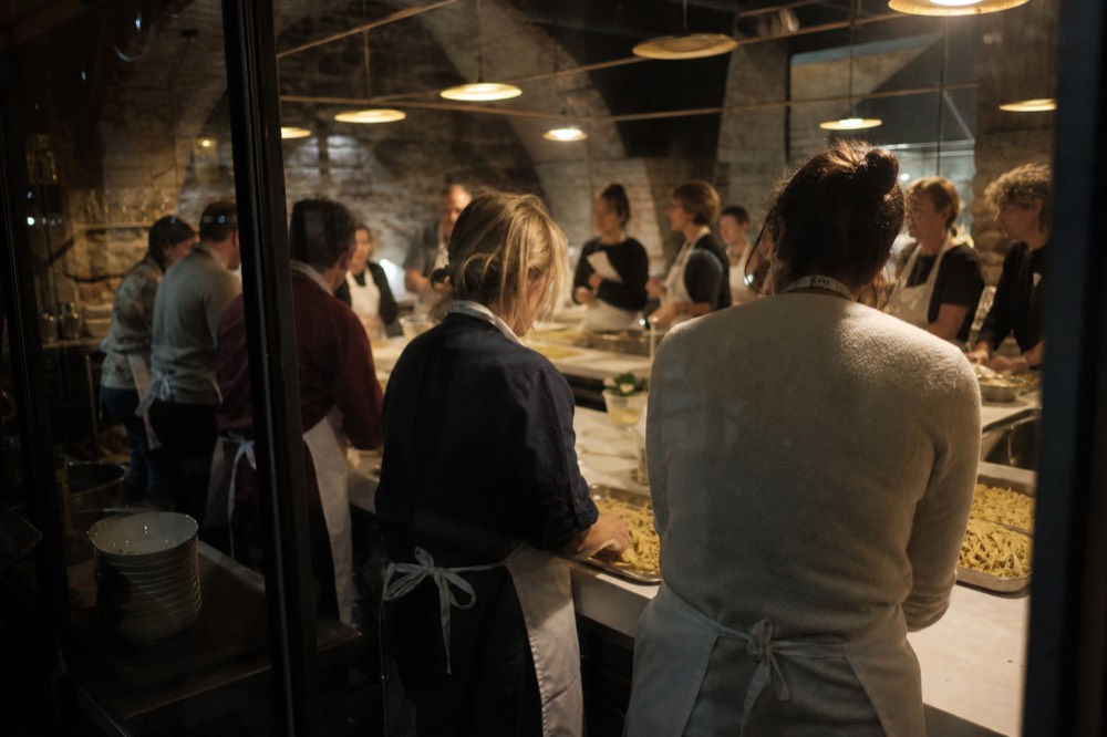 Crowded evening class in a vaulted stone kitchen, a dozen guests laughing over trays of freshly cut tagliatelle under warm hanging lights