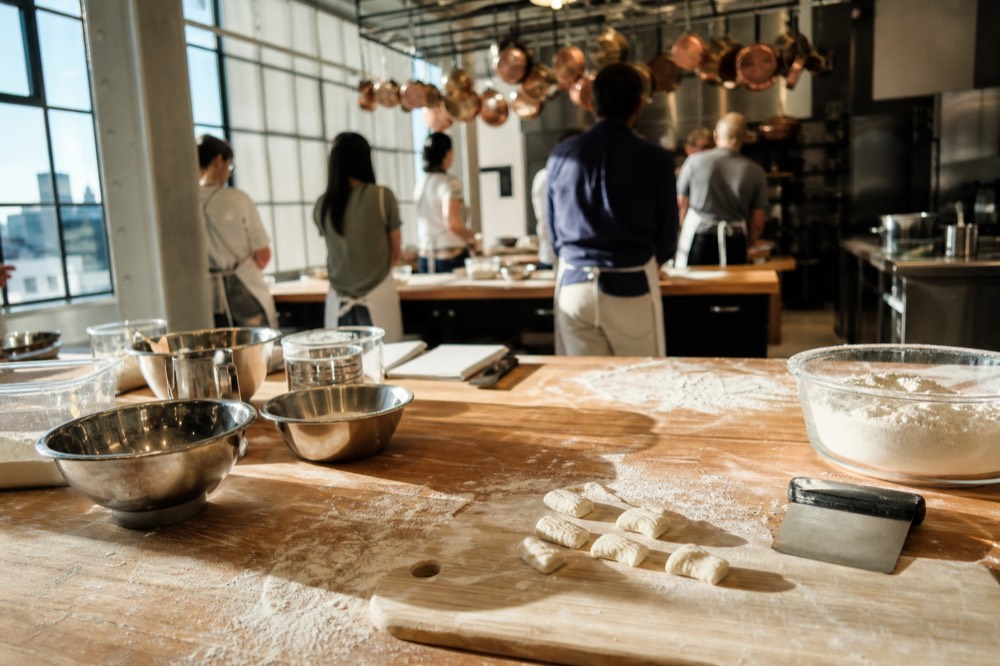 Bright cooking school kitchen, long wooden table, flour-dusted stations, hanging copper pans