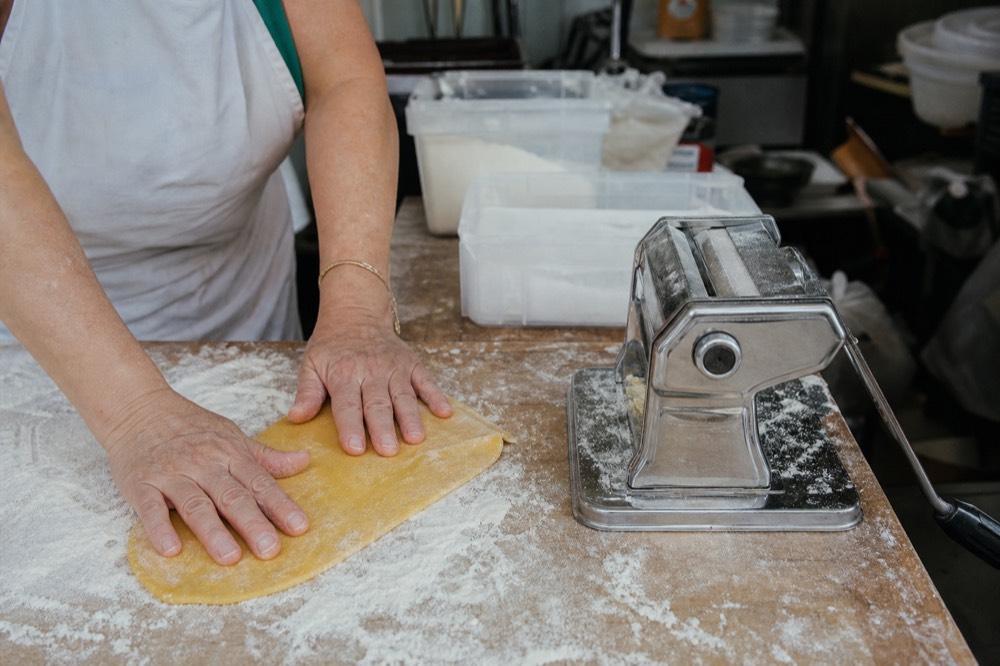 Hands pressing yellow egg dough on a floured board beside a brass pasta cutter
