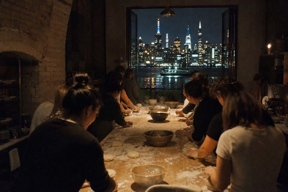 Long communal table in a stone-walled kitchen where a dozen people work dough side by side, flour dust hanging in warm lamplight