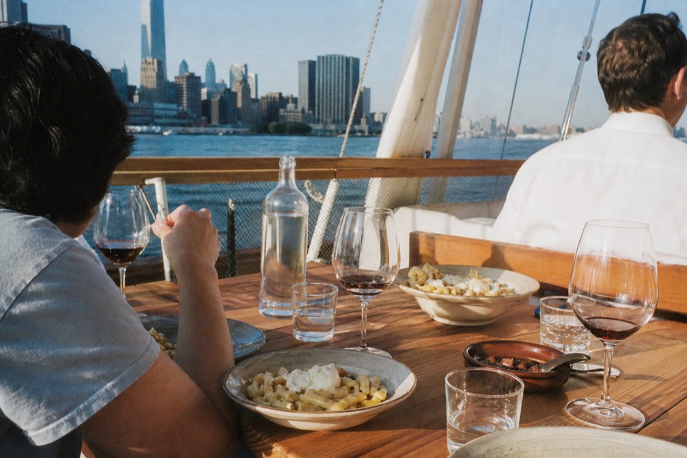 Group of adults seated at a long wooden table with plates of fresh pasta and glasses of red wine, warm evening light