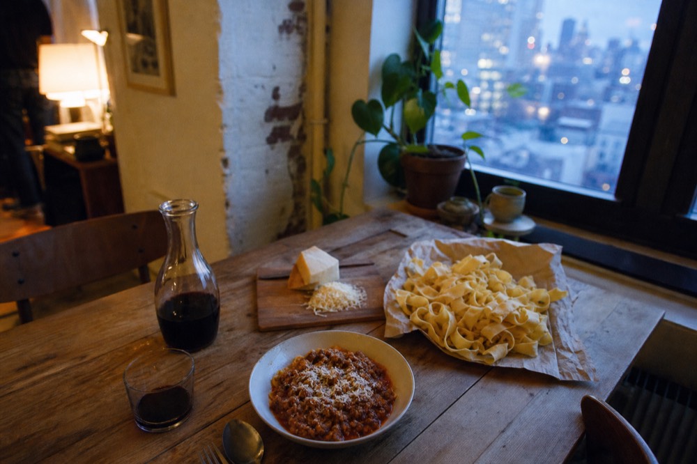 Overhead view of a rustic table set with a bowl of ragu, a carafe of red wine, a wedge of parmesan and a heap of uncooked pappardelle