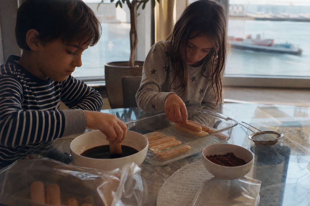 Two children dipping ladyfinger biscuits into a bowl of coffee and layering them into a glass dish, cocoa powder and a sieve waiting beside them