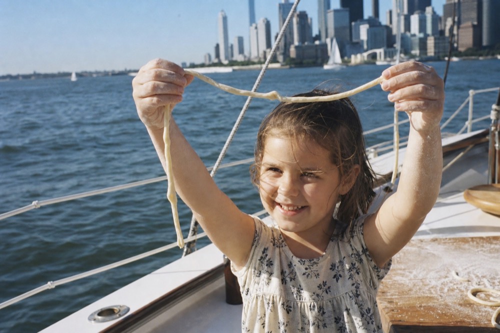 A small girl holding up a single uneven strand of hand-rolled pici with both hands, grinning, floured bench blurred behind her
