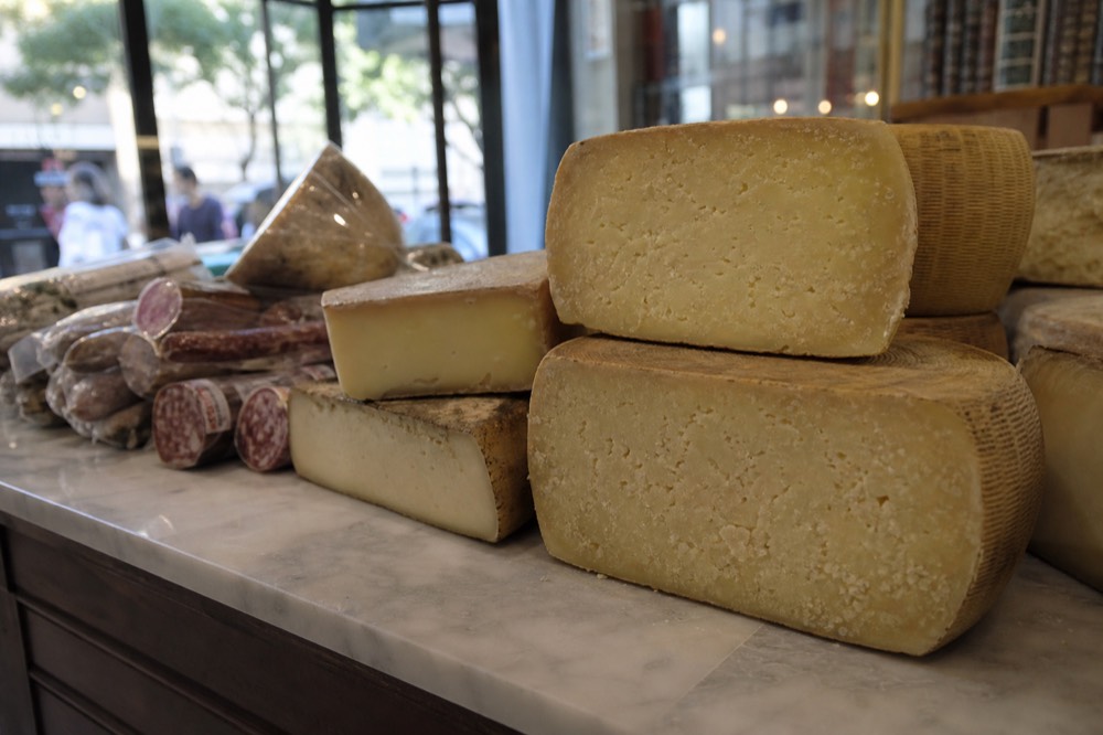 Close view of a market cheese counter, wheels of aged pecorino cut open beside cured meats on a marble surface
