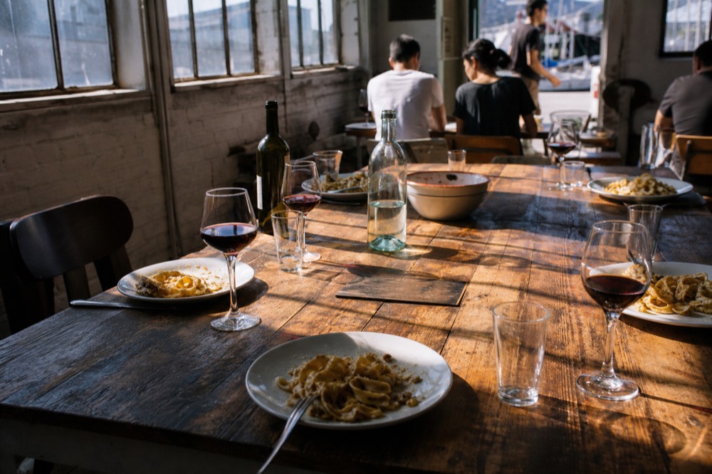 Long table set with plates of fresh pasta and glasses of red wine, sunlight across a rustic dining room after a class