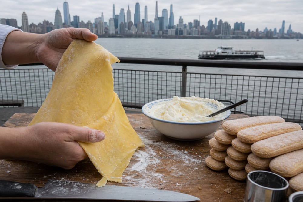 Two hands lifting a sheet of pasta dough off a wooden board while a bowl of whipped mascarpone waits beside a stack of savoiardi