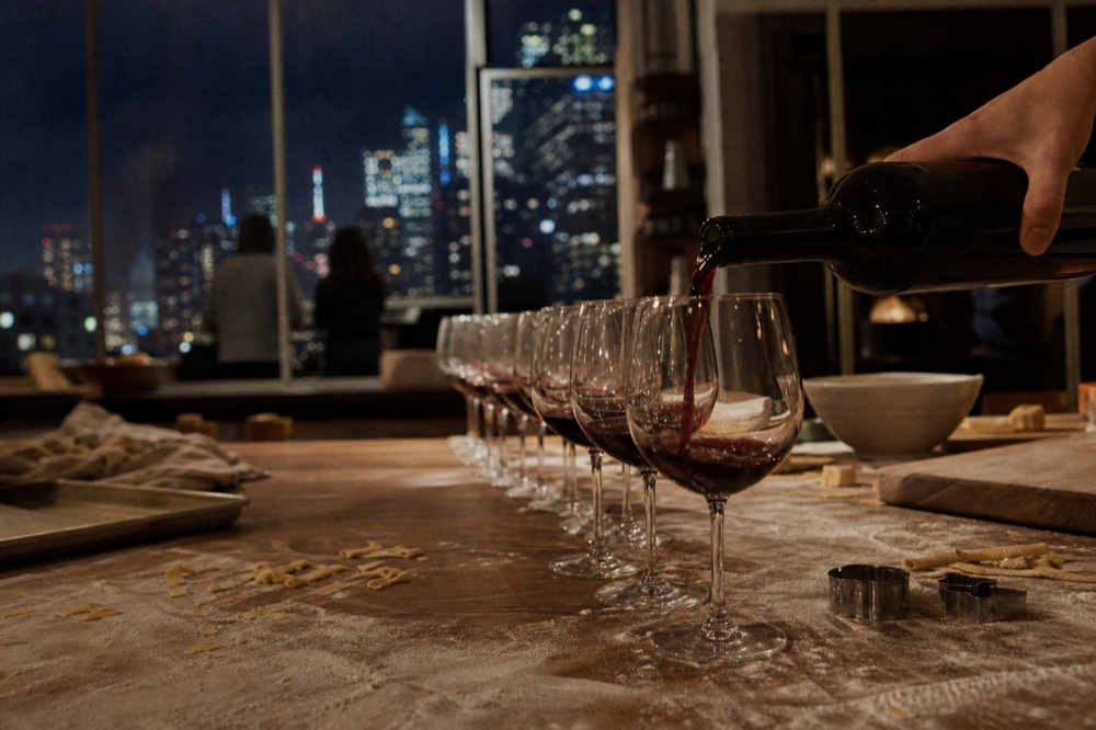 Row of wine glasses being filled with red wine at a long kitchen table scattered with flour and pasta cutters