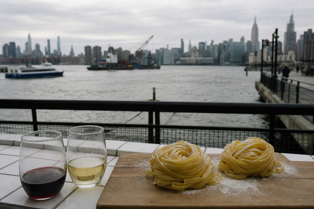 Two nests of fresh tagliatelle dusted with semolina on a wooden board beside a glass of red wine and a glass of white on a tiled counter